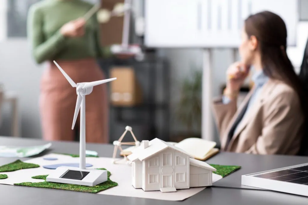 Indonesia Green Building Movement: A model wind turbine and house on a table, with a woman presenting in the background, discussing renewable energy solutions.