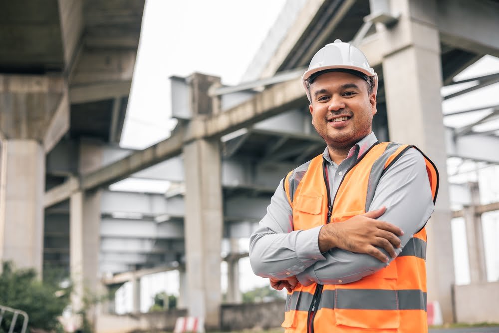 Indonesian engineer handsome man or architect standing confidently in front of a construction site with white safety helmet. Standing at highway concrete road site.