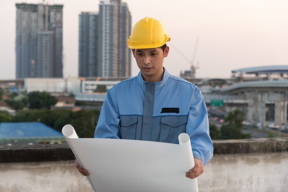 Construction worker in hard hat reading Construction Regulations Indonesia, holding blueprint with cityscape behind.