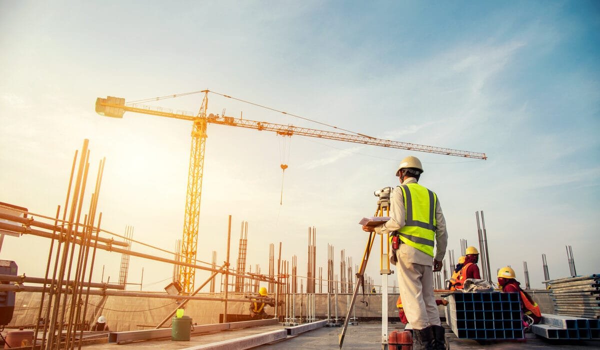 Engineer with blueprint and surveyor tool at construction site checking Construction Regulations Indonesia; workers and crane in background.