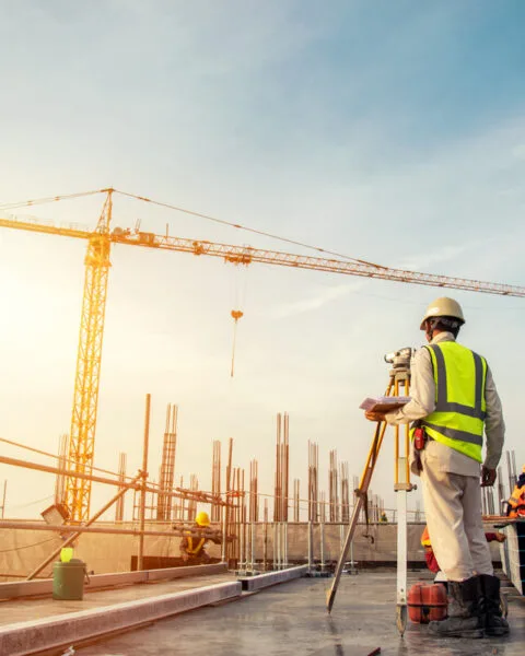 Engineer with blueprint and surveyor tool at construction site checking Construction Regulations Indonesia; workers and crane in background.