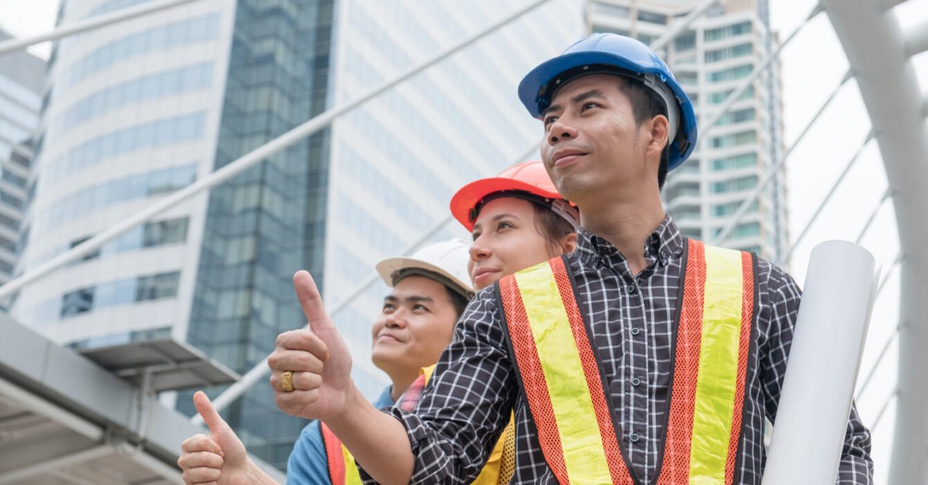 Construction workers in hard hats and safety vests with thumbs up, holding blueprints, in an urban setting, representing Indonesia’s Construction Workforce.