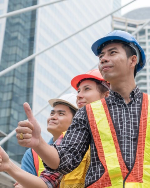 Construction workers in hard hats and safety vests with thumbs up, holding blueprints, in an urban setting, representing Indonesia’s Construction Workforce.