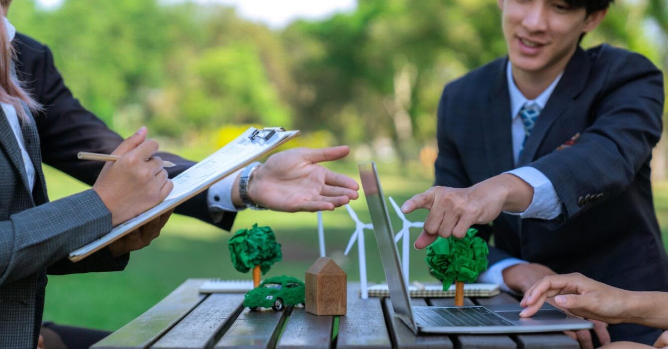 Professionals discussing over a laptop with model wind turbines and a green car on a table, showing Sustainable Infrastructure Projects Indonesia.